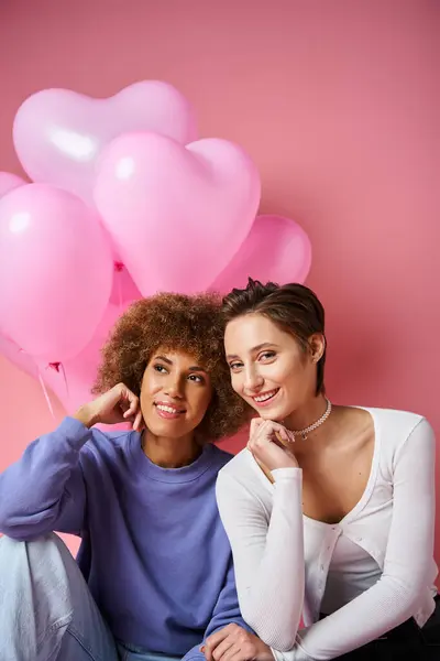 Allegra coppia lesbica multiculturale seduta vicino a palloncini a forma di cuore rosa, San Valentino — Foto stock