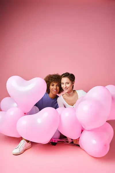 Pareja lesbiana multicultural positiva sentada cerca de globos en forma de corazón rosa, día de San Valentín - foto de stock