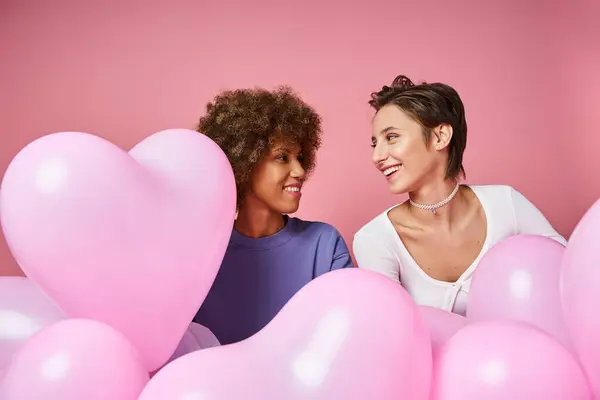 Feliz pareja lesbiana multicultural mirándose cerca de globos en forma de corazón, día de San Valentín - foto de stock