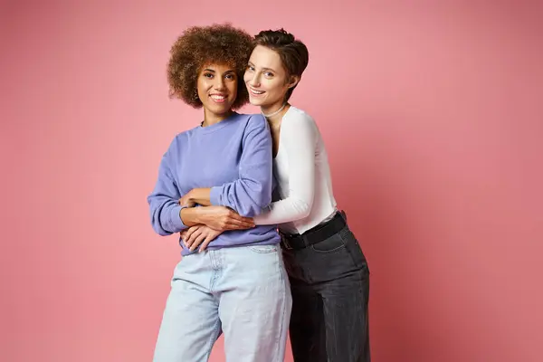 Fröhliche lesbische Frau umarmt afrikanisch-amerikanische Freundin in lässiger Kleidung auf rosa Hintergrund — Stockfoto
