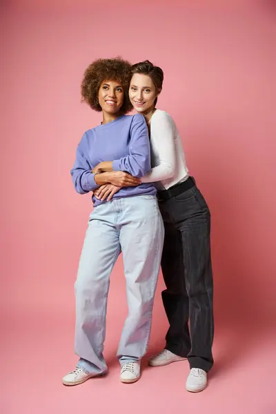 Mujer lesbiana positiva abrazando novia afroamericana en ropa casual sobre fondo rosa - foto de stock