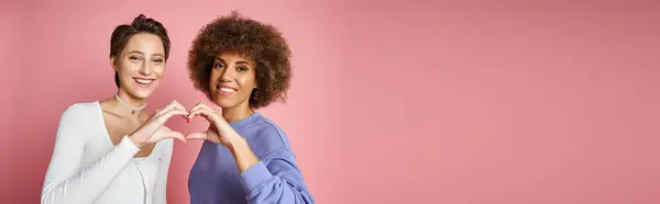 Happy multicultural lesbian couple showing heart sign with hands on pink background, lgbtq banner — Stock Photo
