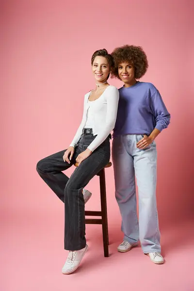 Curly african american woman posing near girlfriend sitting on high stool on pink, lgbtq couple — Stock Photo
