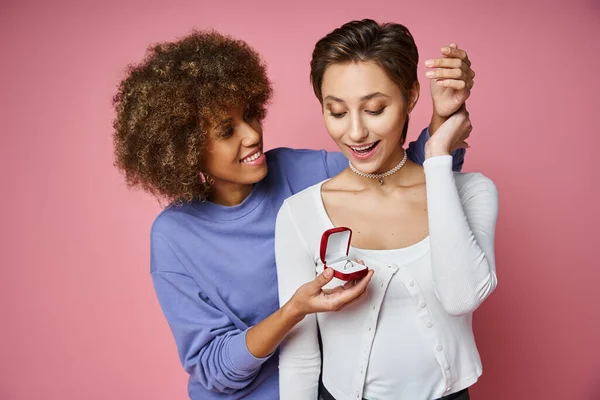 African american woman holding box with engagement ring near excited girlfriend on pink background — Stock Photo