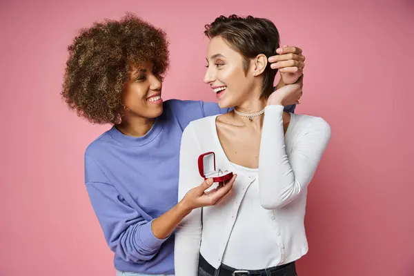 Africaine américaine femme tenant boîte avec bague de fiançailles près de petite amie étonnée sur fond rose — Photo de stock
