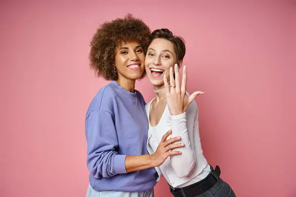 Heureuse lesbienne femme montrant bague de fiançailles sur son doigt près partenaire féminin sur fond rose — Photo de stock