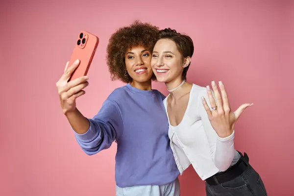 Feliz lesbiana mujer mostrando anillo de compromiso en su dedo mientras toma selfie con prometida en rosa - foto de stock