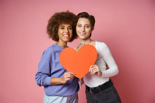 Feliz pareja lesbiana y multiétnica sosteniendo corazón de cartón rojo mientras posan en rosa, día de San Valentín - foto de stock