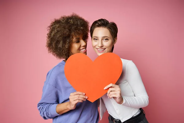 Fröhliches lesbisches und multiethnisches Paar mit rotem Kartonherz, während es am Valentinstag in rosa posiert — Stockfoto