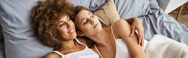 Jeune couple lesbien et multiethnique en pyjama couché ensemble sur le lit à la maison, lgbtq love banner — Photo de stock
