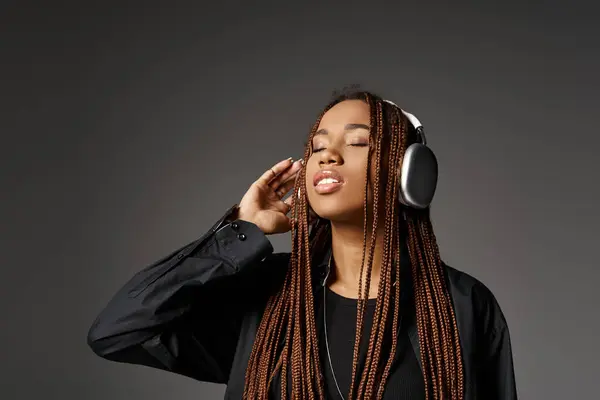 Dreamy african american girl in 20s with dreadlocks listening music in headphones on grey backdrop — Stock Photo