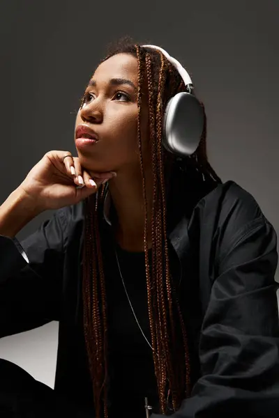 Pensive african american woman in 20s with dreadlocks listening music in stylish headphones on grey — Stock Photo