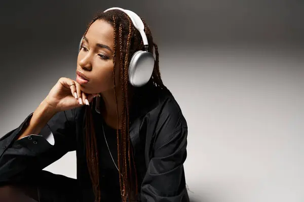Pensive african american woman in 20s with dreadlocks listening music in headphones in grey studio — Stock Photo