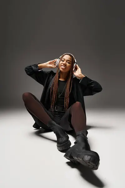 Smiling african american woman in 20s with dreadlocks listening music in headphones in grey studio — Stock Photo