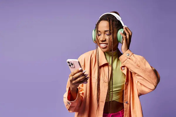 Happy african american woman listening music in her headphones and using phone on lilac backdrop — Stock Photo
