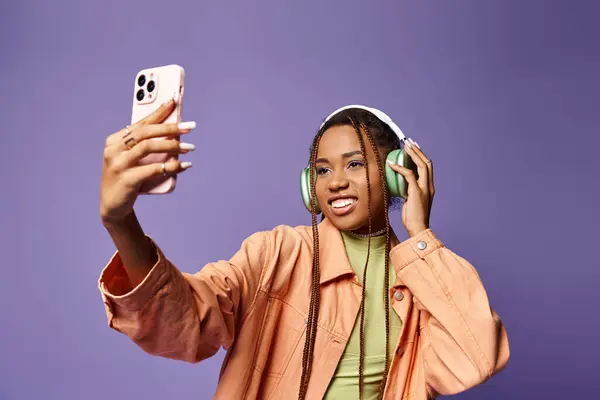 Mulher americana africana alegre na década de 20 tirando selfie com seus fones de ouvido em pano de fundo lilás — Fotografia de Stock