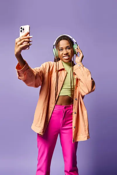 Happy african american woman in 20s taking selfie in her wireless headphones on lilac backdrop — Stock Photo
