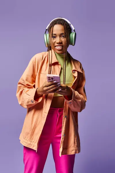 Happy african american woman in green wireless headphones using smartphone on lilac backdrop — Stock Photo