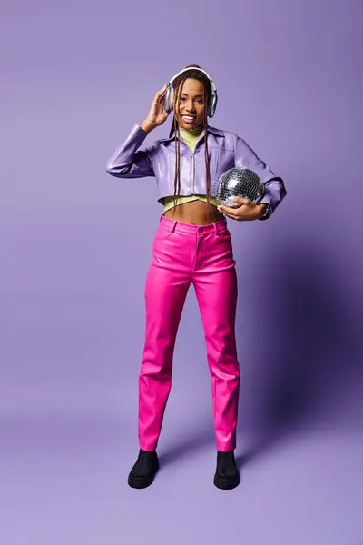 Glückliche junge afrikanisch-amerikanische Frau in stylischer Kleidung und Kopfhörer mit Discokugel auf lila — Stockfoto