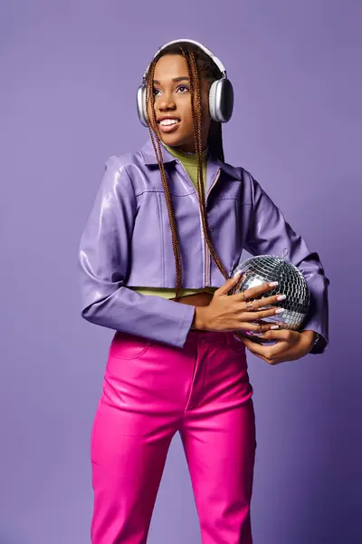Sonriente joven afroamericana mujer en traje elegante y auriculares sosteniendo bola disco en púrpura - foto de stock
