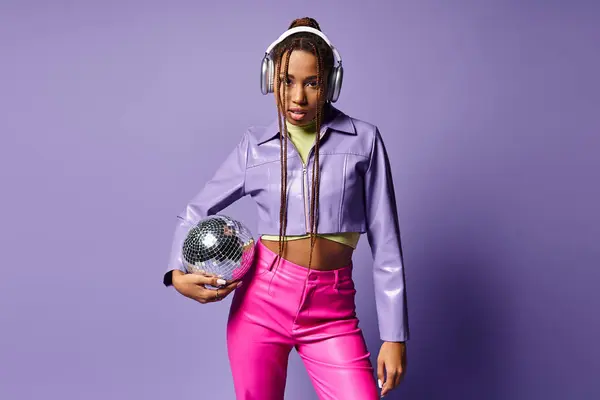 Joven afroamericana mujer en traje de moda y auriculares sosteniendo bola disco sobre fondo púrpura - foto de stock