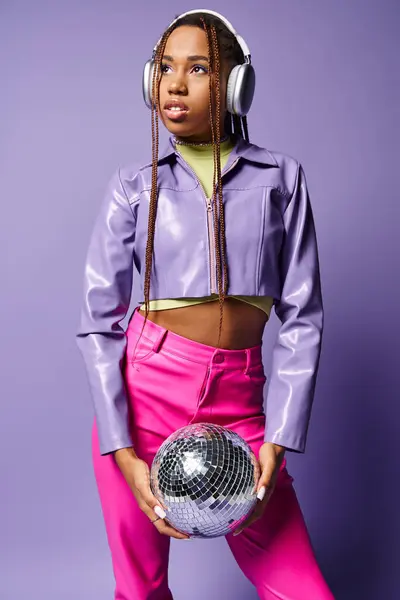 Young african american woman in trendy wireless headphones holding disco ball on purple backdrop — Stock Photo