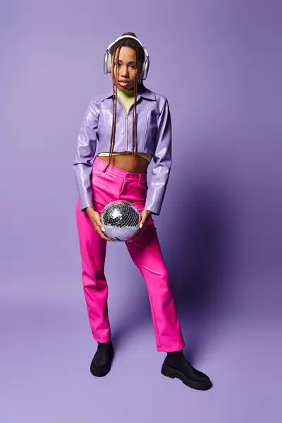 Young african american woman in stylish wireless headphones holding disco ball on purple backdrop — Stock Photo