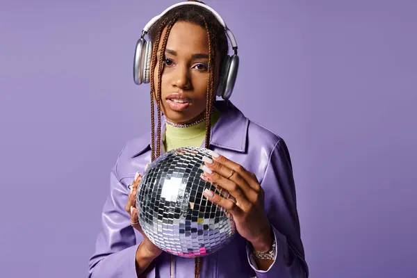 Young african american girl in stylish wireless headphones holding disco ball on purple backdrop — Stock Photo