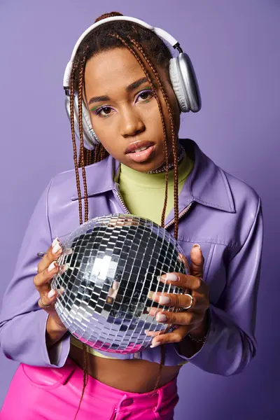 Stylish african american girl in trendy wireless headphones holding disco ball on purple backdrop — Stock Photo