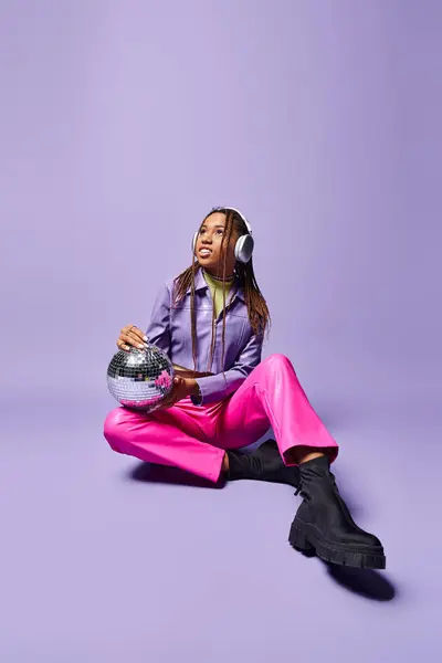 Joyeuse fille afro-américaine en tenue élégante et écouteurs assis avec boule disco sur violet — Photo de stock