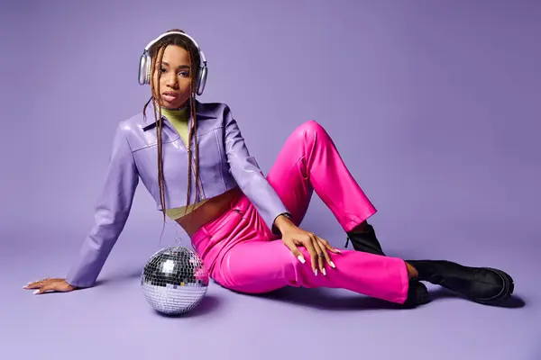 Stylish african american girl in wireless headphones sitting near disco ball on purple backdrop — Stock Photo