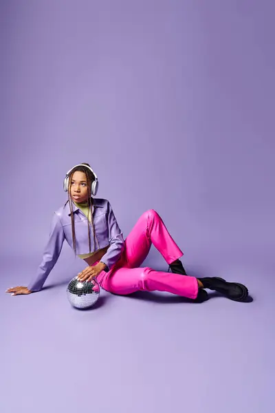 Menina americana africana na moda em fones de ouvido sem fio sentado perto de bola de discoteca em pano de fundo roxo — Fotografia de Stock