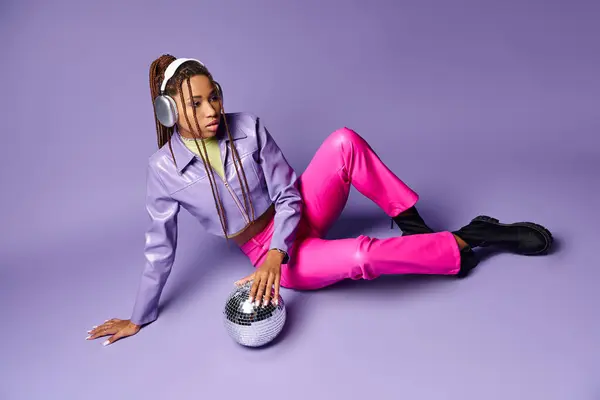 Donna afroamericana in cuffie wireless alla moda seduta vicino alla palla disco su sfondo viola — Foto stock