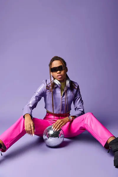 Mulher americana africana na moda óculos de sol e fones de ouvido sentado perto de bola de discoteca em roxo — Fotografia de Stock