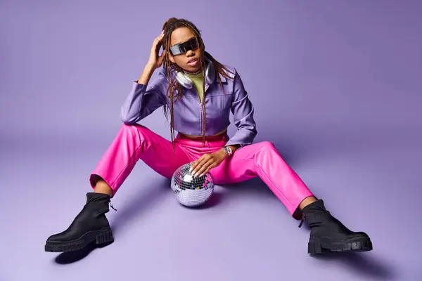 African american girl in trendy sunglasses and headphones sitting near disco ball on purple — Stock Photo