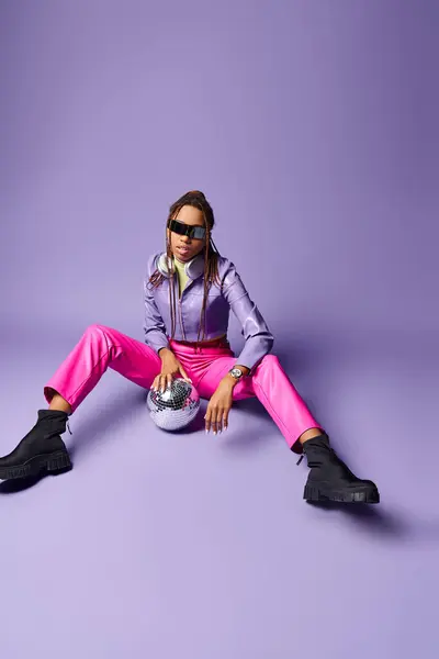 Stylish african american girl in trendy sunglasses and headphones sitting near disco ball on purple — Stock Photo