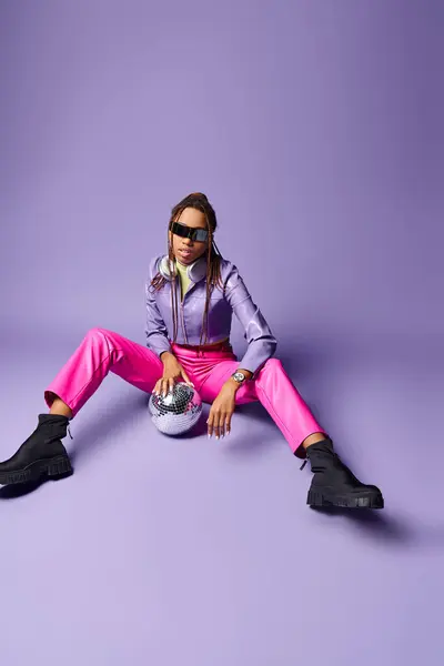 Chica afroamericana con estilo en gafas de sol de moda y auriculares sentados cerca de la bola disco en púrpura - foto de stock