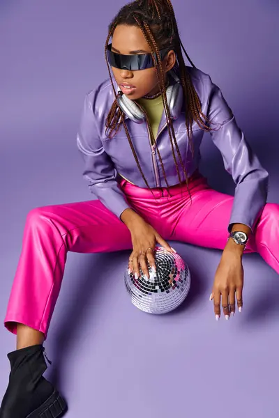 Young african american woman in trendy sunglasses and headphones sitting near disco ball on purple — Stock Photo