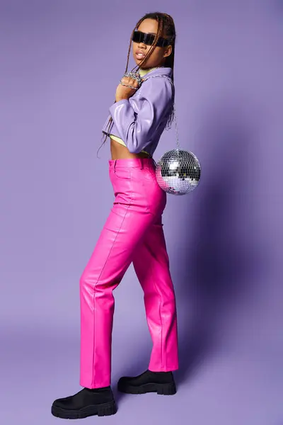 Афроамериканська дівчина в футуристичних сонцезахисних окулярах тримає диско-кулю на ланцюжку і стоїть на фіолетовому — стокове фото