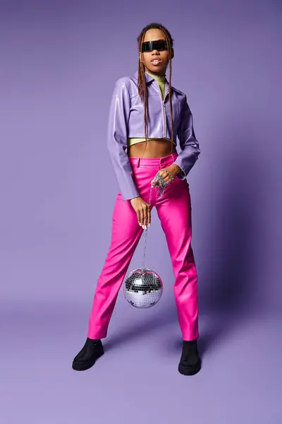 African american woman in trendy sunglasses holding disco ball on chain and standing on purple — Stock Photo