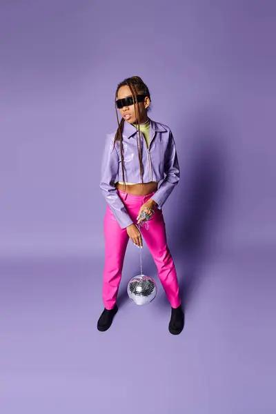 Afro-américaine dans des lunettes de soleil élégantes tenant boule disco sur la chaîne et debout sur violet — Photo de stock