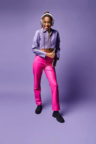 Mujer afroamericana feliz con auriculares que sostienen el ordenador portátil y de pie sobre fondo púrpura - foto de stock