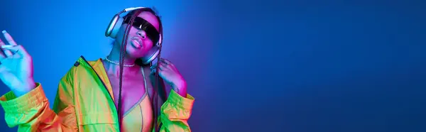 Chica afroamericana en auriculares y gafas de sol escuchando música en estudio con luces, pancarta - foto de stock