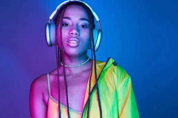 Beautiful dark-skinned woman in wireless headphones posing in jacket in studio with neon lights — Stock Photo