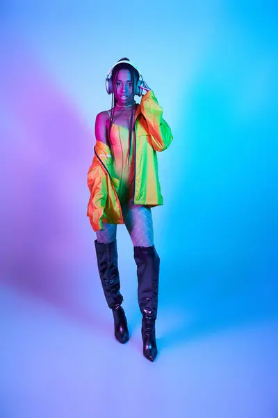 Темнокожая девушка в наушниках, позирующая в телесном костюме и на коленных сапогах в студии с неоновыми огнями — стоковое фото