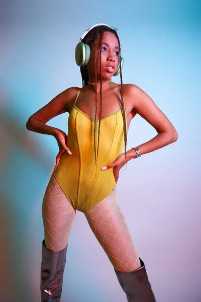 Joven mujer afroamericana en auriculares posando en traje de cuerpo amarillo y botas de rodilla en estudio - foto de stock