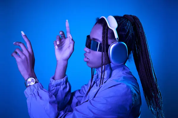Dreamy african american woman posing in trendy sunglasses with headphones on blue with neon light — Stock Photo
