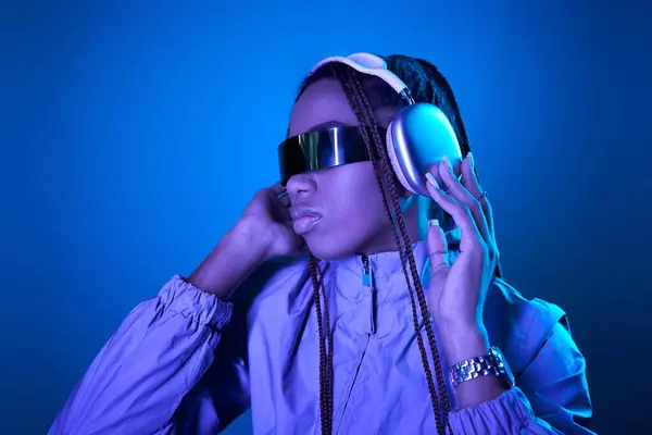 Pretty african american woman posing in trendy sunglasses with headphones on blue with neon light — Stock Photo