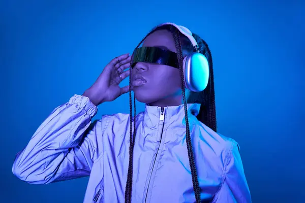 Pretty dark skinned woman posing in trendy sunglasses with headphones on blue with neon light — Stock Photo