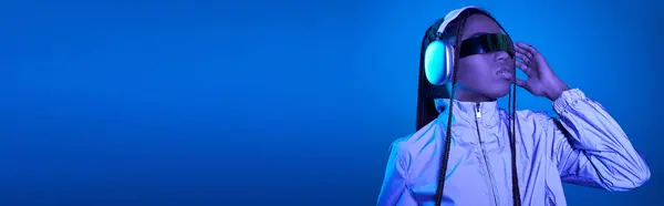 Pretty dark skinned girl posing in trendy sunglasses with headphones on blue with neon light, banner — Stock Photo